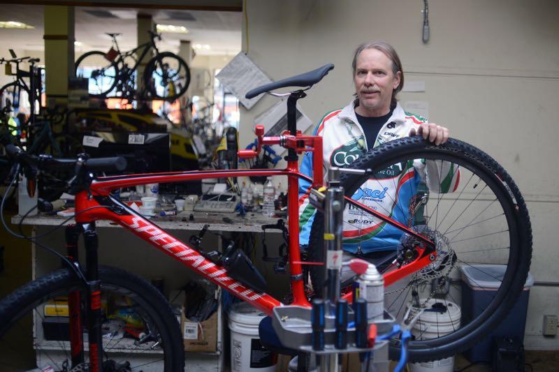A man in a cycling jersey stands beside a red mountain bike, holding a rear wheel. The bike is on a repair stand in a bike shop, surrounded by tools and cycling equipment. In the background, various bicycles are displayed.