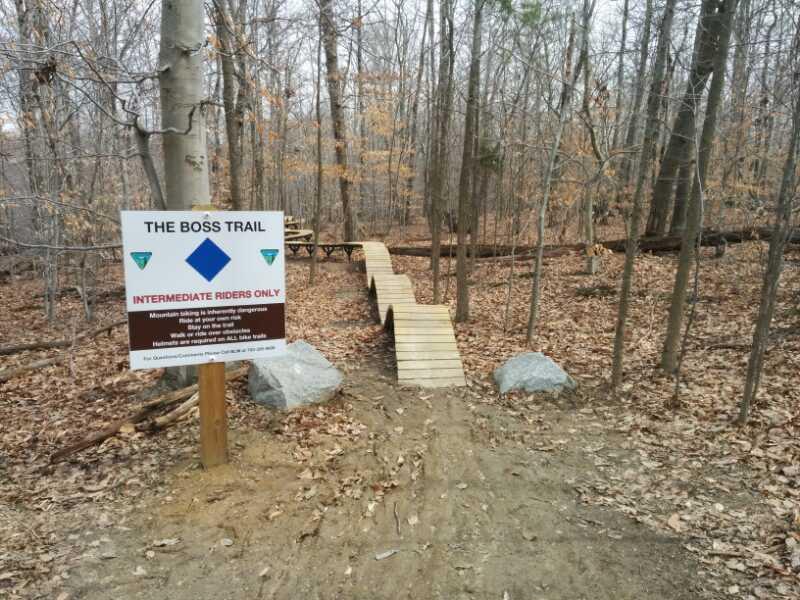 A dirt path through a wooded area with a wooden bridge designed for mountain biking. A sign beside the path reads "The Boss Trail," indicating it is for intermediate riders only. The surrounding trees have no leaves, suggesting it is autumn or early spring. Meadowood mountain bike trail.
