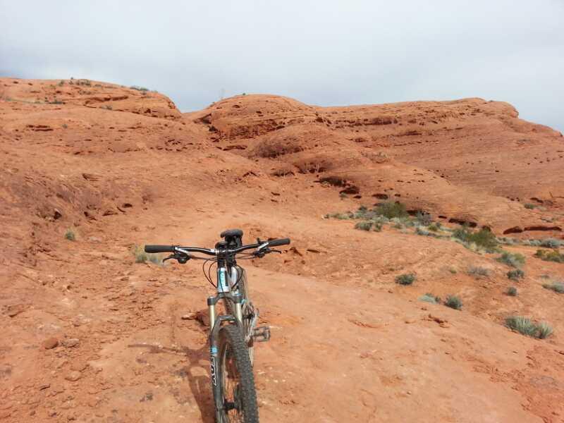 Mountain bike positioned on a rocky, red dirt trail with rugged terrain and a hilly landscape in the background, under a cloudy sky. Prospector - Church Rocks Loop mountain bike trail.
