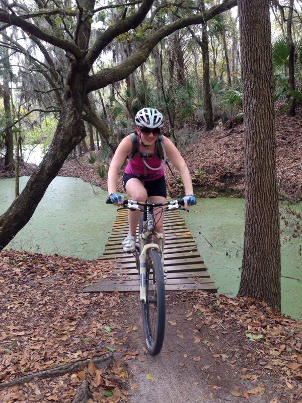 A woman in a pink tank top and shorts rides a mountain bike over a wooden bridge surrounded by trees and a pond covered with green vegetation. The scene is set in a wooded area with fallen leaves on the ground. Alafia River State Park mountain bike trail.