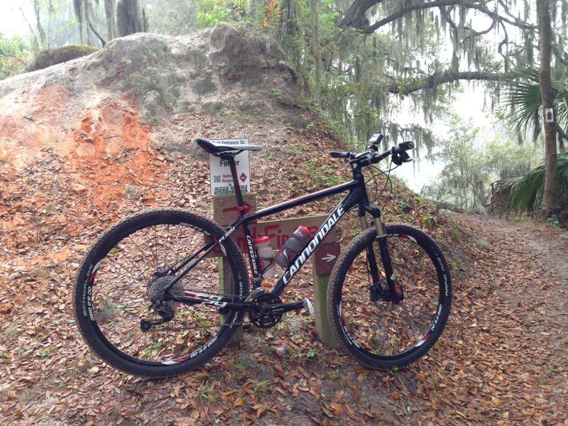 A mountain bike leaning against a trail sign on a dirt path surrounded by fall leaves and greenery, with a rocky background and misty atmosphere. The bike features black and red colors with a "Cannondale" logo. Loyce E. Harpe Park mountain bike trail.