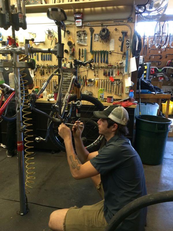 A mechanic working on a bicycle in a workshop. The bike is mounted on a repair stand, and various tools and equipment are visible on a pegboard in the background. The mechanic is focused on performing maintenance, using a tool while sitting on the floor. The workspace is well-organized with additional bike parts and a trash can in the vicinity.