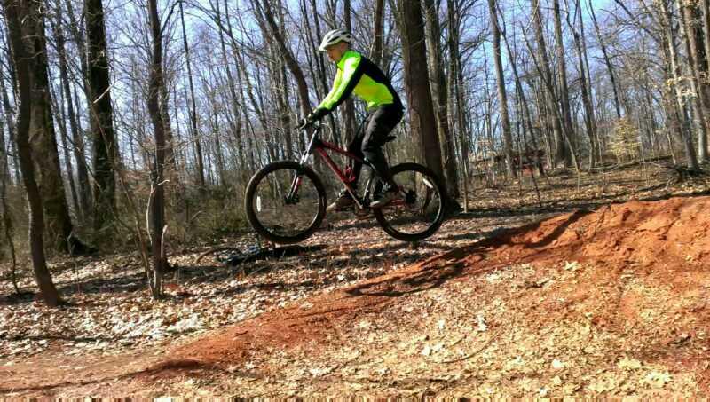 A mountain biker in a bright neon green jacket and helmet is mid-air as they jump off a dirt ramp in a forested area. The ground is covered in fallen leaves, and trees surround the scene, creating a natural backdrop. Freedom Center mountain bike trail.