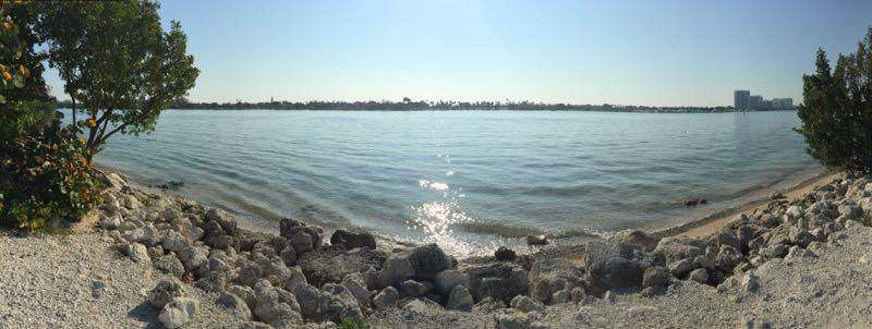 A panoramic view of a calm waterfront with gentle waves, surrounded by rocky shoreline and greenery. The scene features clear blue skies and the sun reflecting off the water, with a distant view of buildings along the shoreline. Oleta River State Park mountain bike trail.