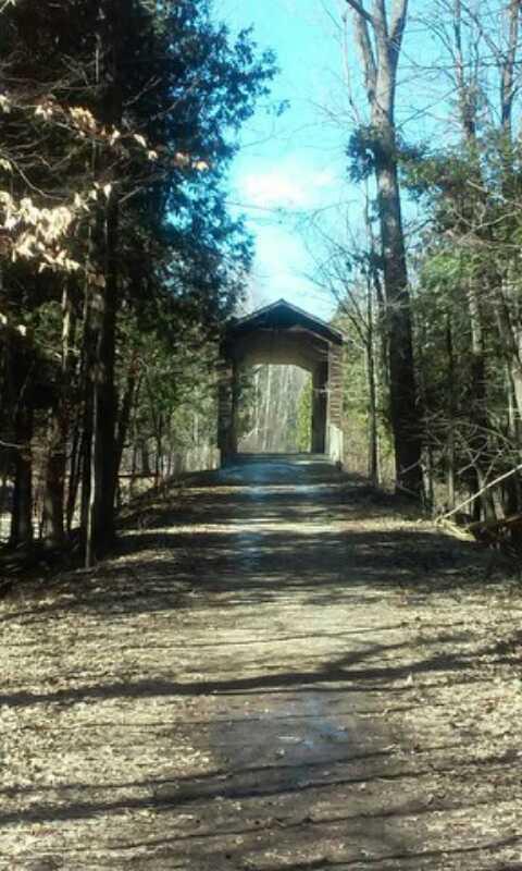 A dirt path leads through a tranquil forest, lined with tall trees and scattered with fallen leaves. In the distance, a wooden archway marks the entrance to a covered bridge, illuminated by soft sunlight filtering through the branches above. The scene conveys a sense of peacefulness and natural beauty. Deerfield Park mountain bike trail.
