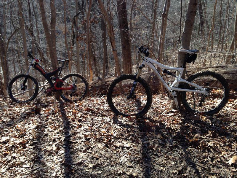 Two mountain bikes are parked side by side on a forest trail covered with dry leaves. The bike on the left is primarily black with red accents, while the bike on the right is white with black details. Surrounding them are tall, bare trees in a wooded area, creating a serene outdoor setting. Wissahickon Valley Park mountain bike trail.