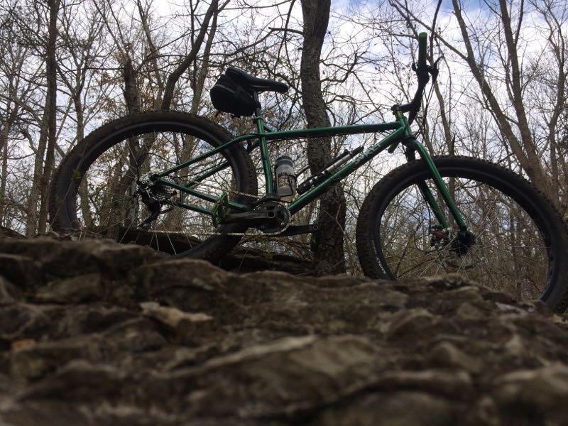 A green mountain bike resting on rocky terrain, surrounded by bare trees under a cloudy sky. The bike is positioned at an angle, showcasing its frame and wheels, with a water bottle attached to the frame. Landahl Park Reserve mountain bike trail.