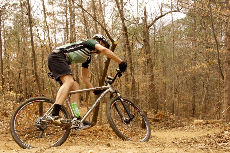 A mountain biker navigating a dirt trail surrounded by bare trees in a forested area. The rider is leaning forward on the bike, wearing a helmet and a green and black cycling jersey. A water bottle is attached to the bike frame. The scene captures the essence of outdoor adventure and mountain biking.
