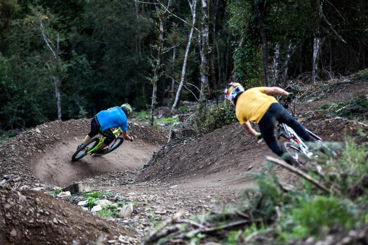 Two mountain bikers navigate a dirt trail in a forested area, leaning into a curve. One rider wears a blue shirt and helmet, while the other has a yellow shirt. The surrounding terrain is uneven, with trees and shrubs visible in the background.