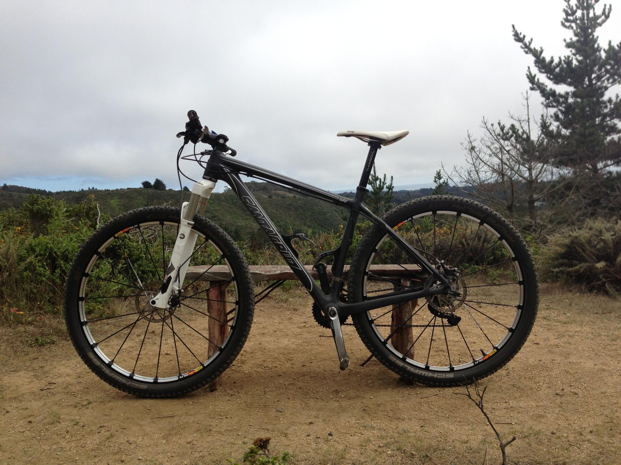 Santa Cruz Highball Carbon: A mountain bike parked on a dirt trail with a scenic backdrop of hills and trees under a cloudy sky. The bike has a black frame and white front suspension fork, with a visible disc brake on the front wheel.