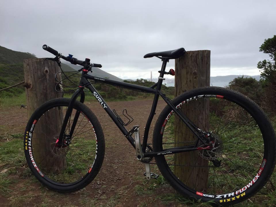 Surly Karate Monkey: A black mountain bike parked next to wooden posts on a gravel path, surrounded by green foliage and hills under a cloudy sky. The bike has distinctive wheels and is facing the camera, showcasing its frame and details.