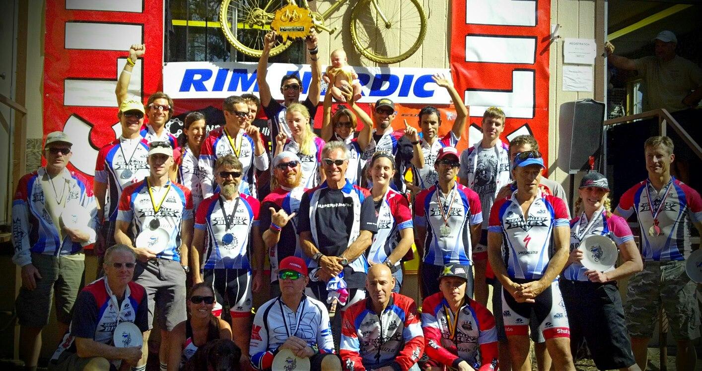 A large group of people posing for a photo in front of a red and white backdrop decorated with a bicycle. Many individuals are wearing matching cycling jerseys and medals, indicating a celebration or award ceremony, possibly after a cycling event. Some participants are holding trophies, while others show enthusiastic gestures. A baby is being held by one person in the center of the group.
