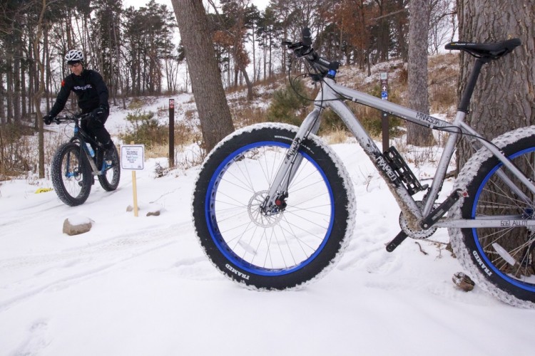 A person riding a fat tire bike on a snow-covered trail, surrounded by trees in a winter landscape. A second fat tire bike with a silver frame and blue rims is parked nearby, with a sign in the background indicating that the trail is open.