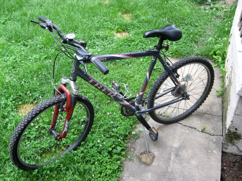 Trek 820: A gray and red mountain bike is leaning against a cement path in a grassy area. The bike features thick tires designed for off-road use and has a comfortable black saddle. The handlebars are positioned at an angle, and the bike's frame is visible with a brand name displayed prominently. There is a patch of green grass and a few small flowers in the background.