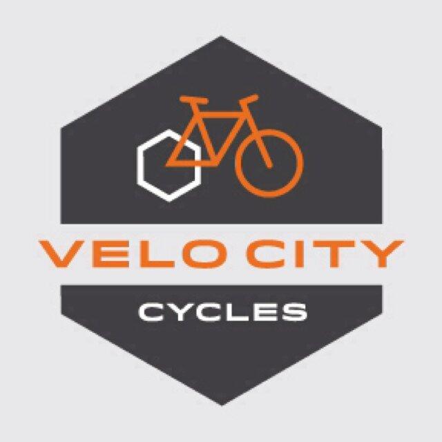 Logo of Velo City Cycles featuring an orange bicycle illustration and the text "VELO CITY" in bold orange letters on a dark hexagonal background, with the word "CYCLES" below in a smaller white font.