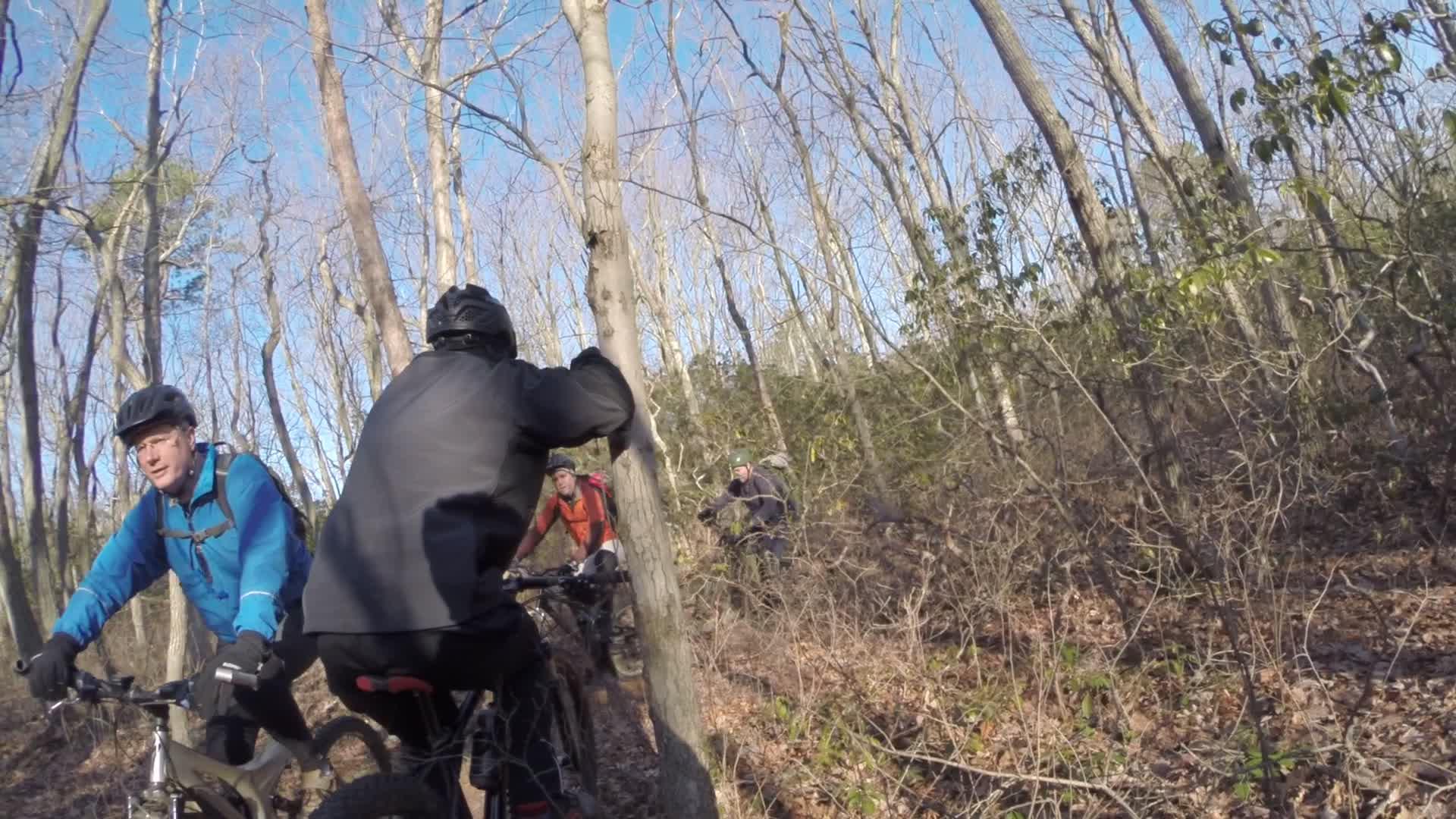 Three mountain bikers navigating through a wooded trail. The scene features bare trees and underbrush in a natural setting. One biker in a blue jacket appears to be looking back, while another wears a black coat, and a third biker, wearing an orange jacket, is partially visible in the background. The sky is clear, indicating good weather for cycling. Allaire State Park mountain bike trail.