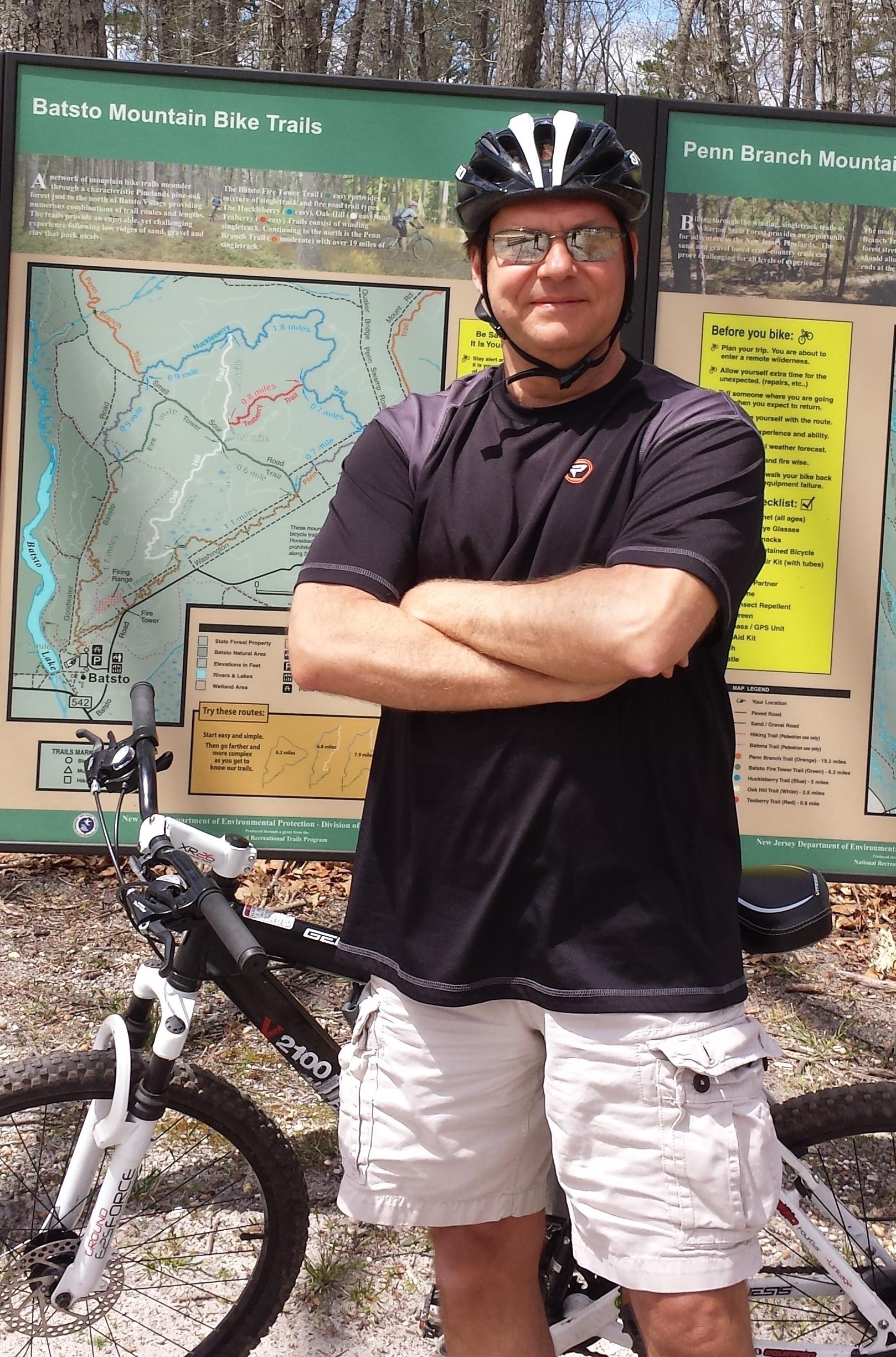 A person in a black cycling shirt and shorts stands confidently with arms crossed next to a mountain bike. They are wearing a helmet and sunglasses. Behind them is a large informational sign detailing the Batsto Mountain Bike Trails, with a map and guidelines for biking. The setting appears to be a wooded area. Wharton State Forest mountain bike trail.