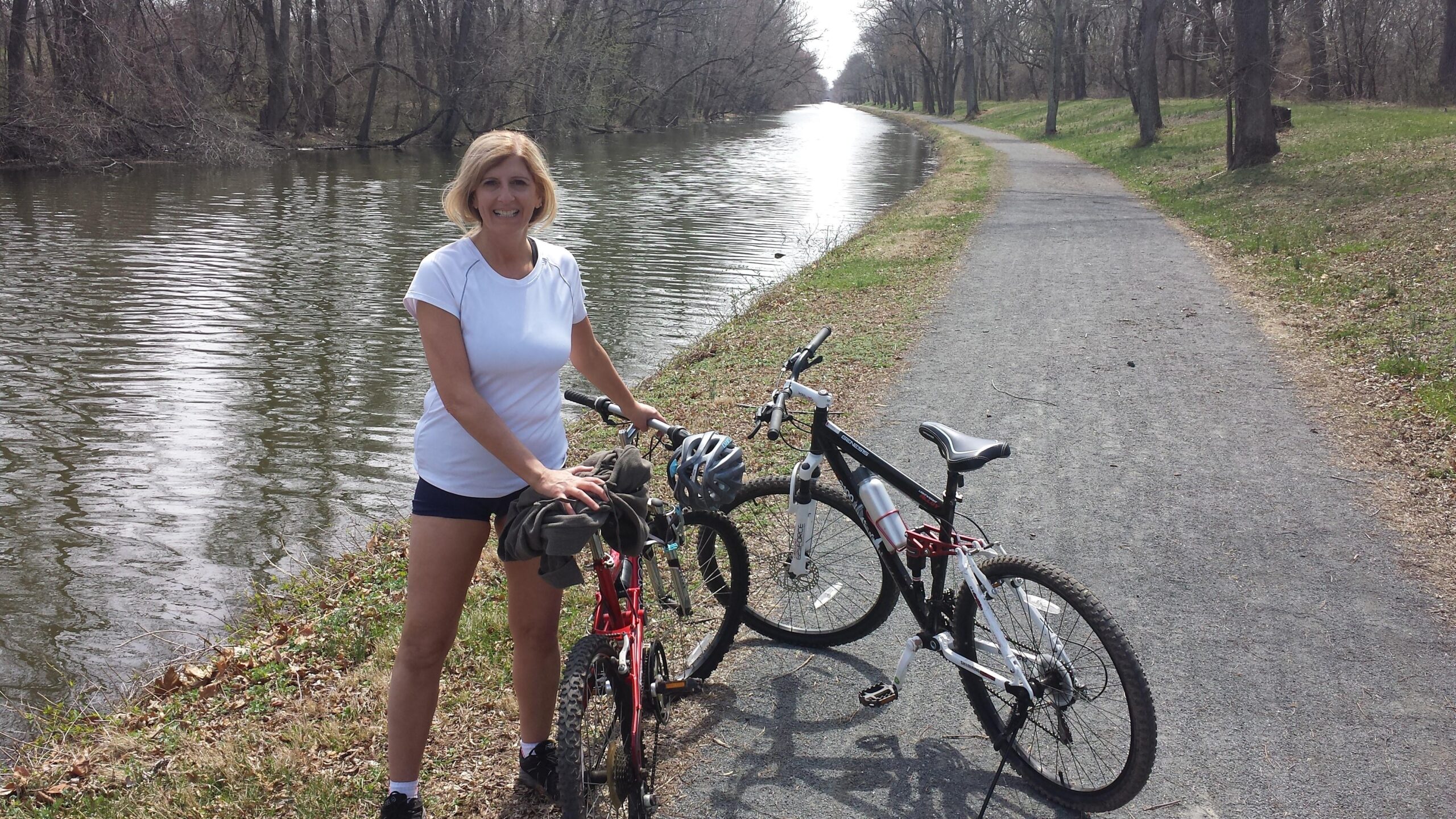 A smiling woman stands beside two bicycles on a gravel path next to a serene canal. She is wearing a white athletic shirt and shorts, and is holding a jacket over the handlebars of the red bike. The surrounding area features bare trees and grass, indicating early spring. The calm water reflects the trees, creating a peaceful outdoor scene. Canal Ride mountain bike trail.