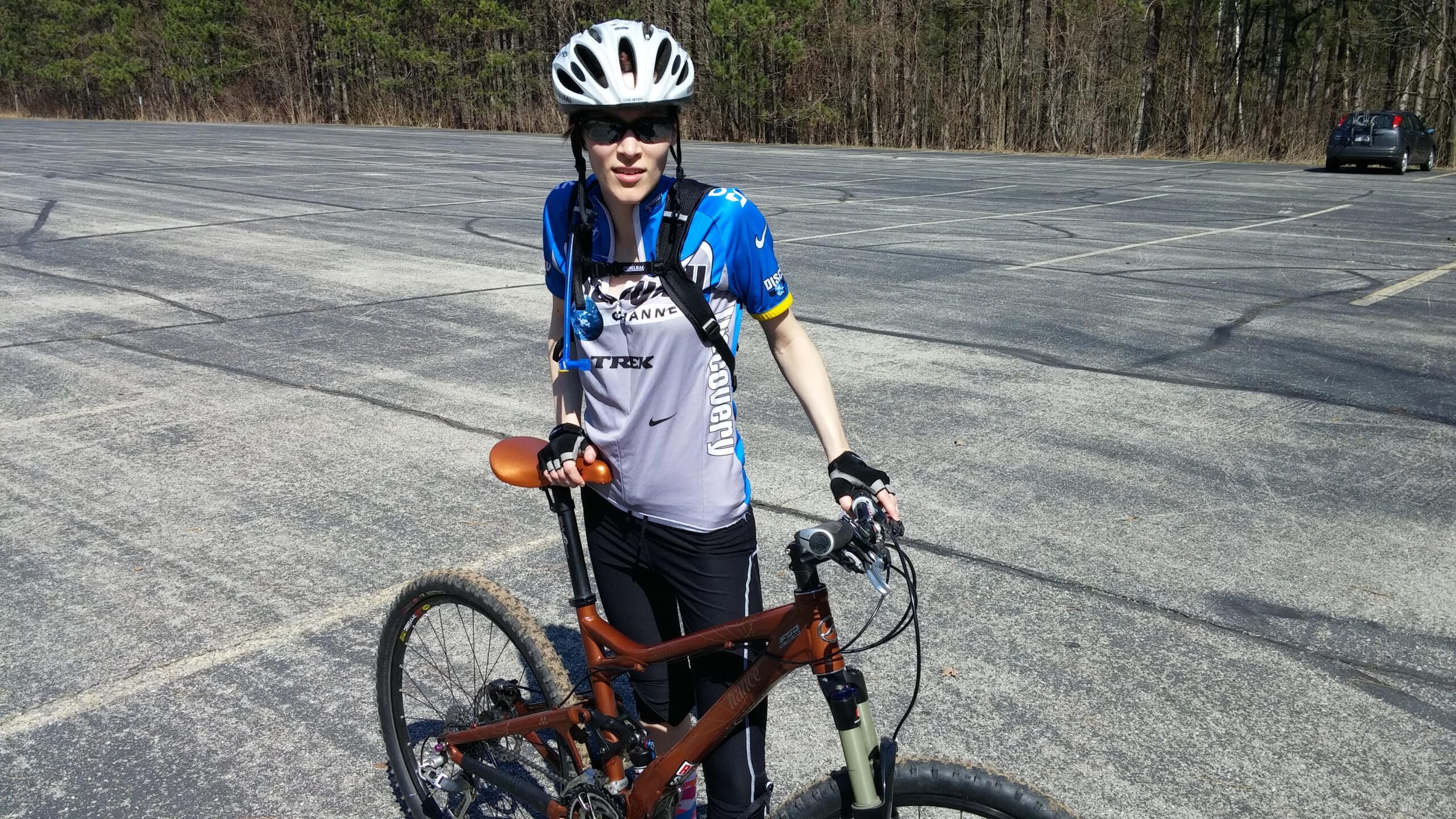 Giant Trance 27.5 sx: A person wearing a cycling helmet, sunglasses, and a blue cycling jersey is standing next to a brown mountain bike in an empty parking lot. The background features trees, indicating a natural setting. The cyclist appears ready for a ride.