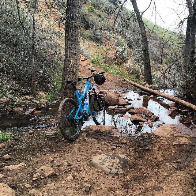 A blue mountain bike resting on a dirt trail near a small stream, surrounded by trees and rocky terrain. Red Rocks / Dakota Ridge mountain bike trail.