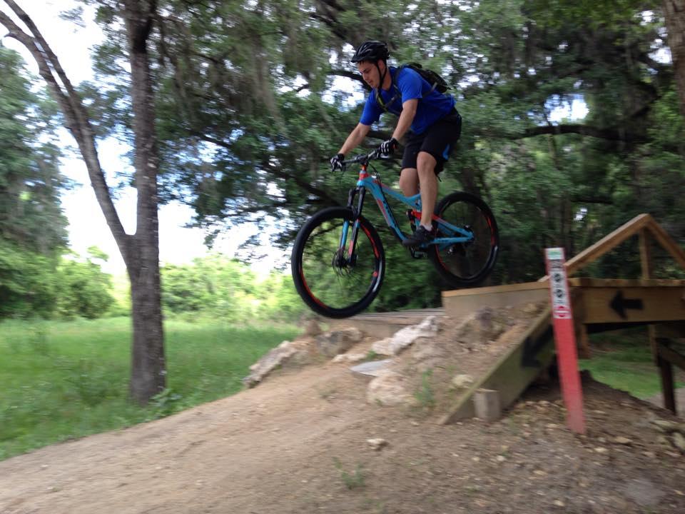 A person in a blue shirt and black shorts rides a mountain bike while jumping off a wooden ramp on a dirt trail surrounded by trees and greenery. Santos mountain bike trail.