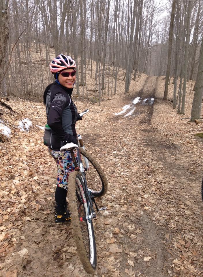 Motobecane NightTrain Bullet: A cyclist in colorful leggings and a black jacket smiles while carrying a mountain bike on a leaf-covered trail. The background features a serene forest with bare trees and patches of snow on the ground, indicating early spring.