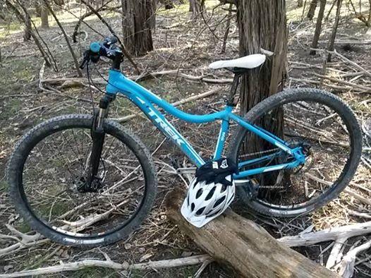 Trek Skye S: A blue mountain bike leaning against a log in a wooded area, with a white helmet placed on the log beside it. The background features trees and scattered branches, indicating a natural outdoor setting.