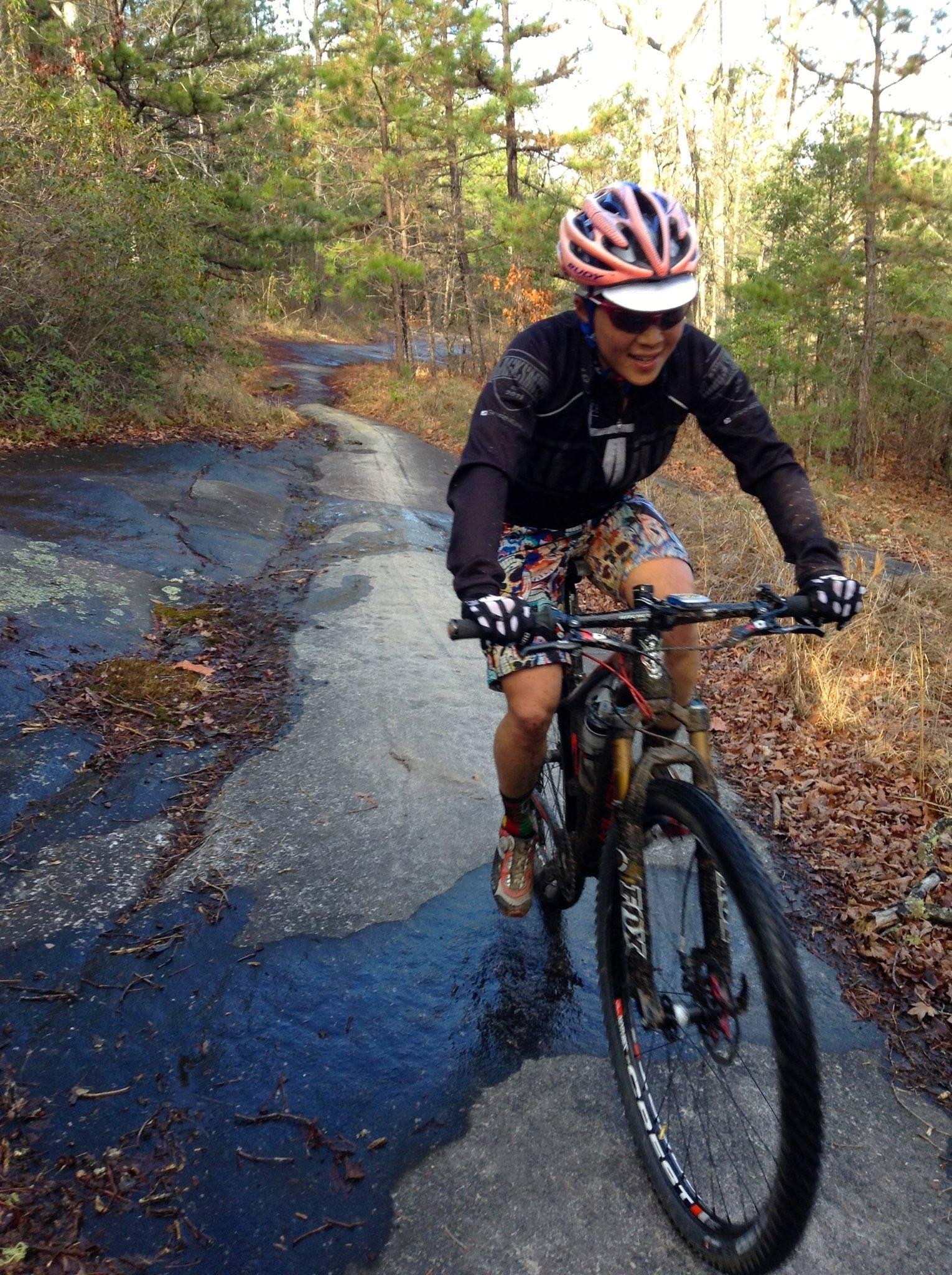 A cyclist navigating a rocky trail surrounded by trees, wearing a helmet and colorful cycling attire. The path shows signs of moisture, and fallen leaves are scattered along the ground. DuPont State Recreational Forest mountain bike trail.