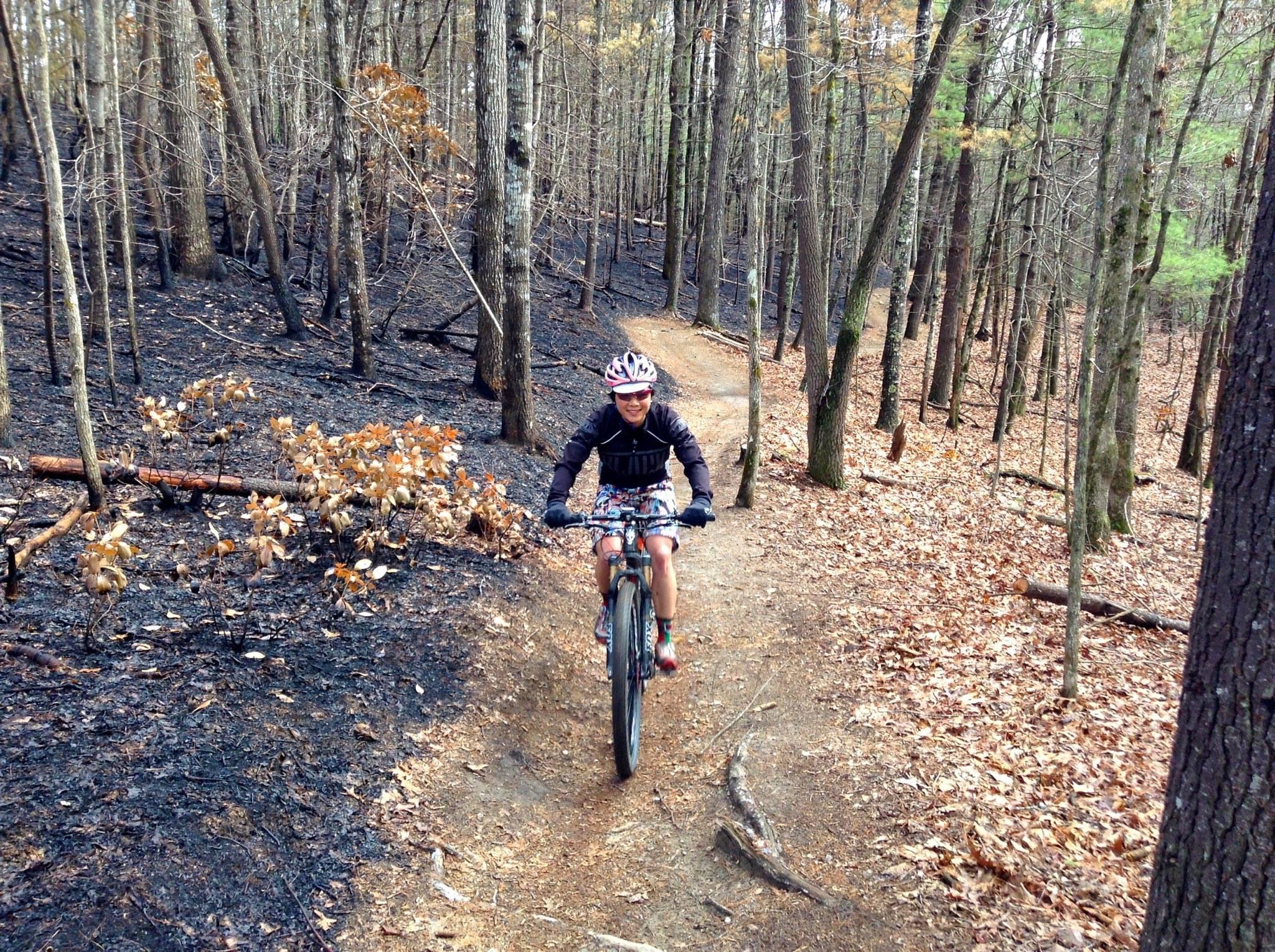A person riding a mountain bike on a narrow dirt trail through a forest that shows signs of recent fire damage, with blackened ground and trees. The rider is wearing a helmet, a black jacket, and colorful shorts, and appears to be smiling as they navigate the trail. DuPont State Forest mountain bike trail.