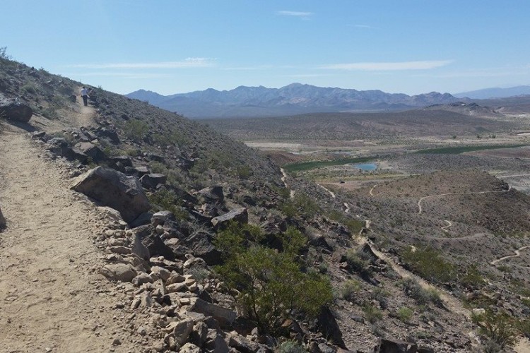 A hiking trail winding along a rocky hillside with sparse vegetation and views of mountains in the background. A small body of water is visible in the valley below, under a clear blue sky.