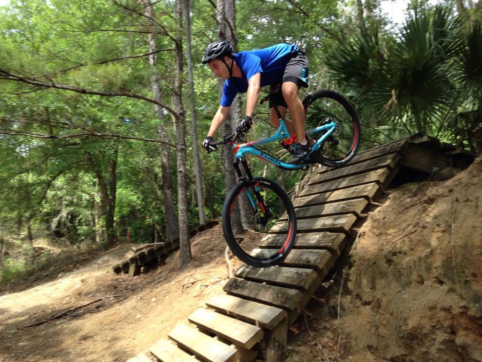A mountain biker in a blue shirt and black shorts jumps off a wooden ramp in a forested area. Trees and greenery surround the path as the cyclist navigates the trail, wearing a helmet and gloves for safety. Santos mountain bike trail.