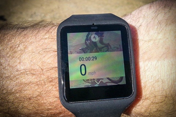 A close-up view of a black smartwatch displaying a timer reading 00:00:29 and a speed of 0 mph on its screen. The watch is worn on a person’s arm, with visible arm hair and a blurred outdoor background.