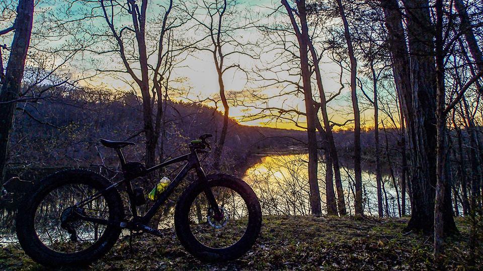 A mountain bike with wide tires is positioned beside a calm river at sunset, surrounded by bare trees. The sky displays a gradient of warm colors, reflecting on the water's surface. Chimney Rock mountain bike trail.