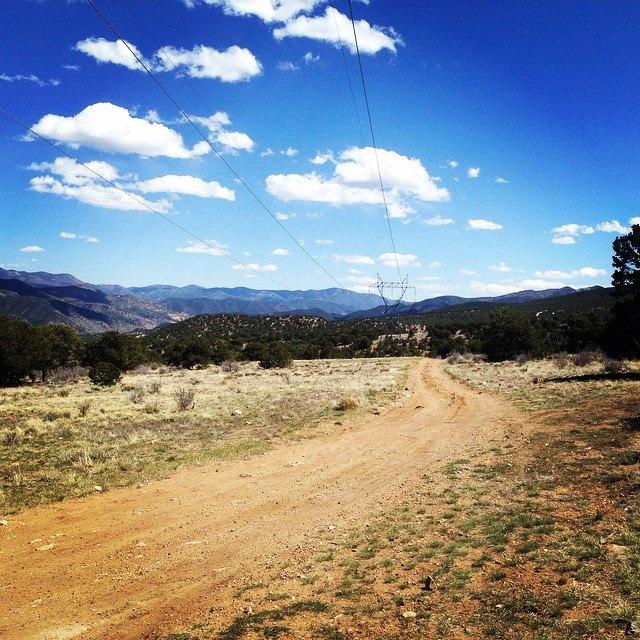 A dirt path winding through an open landscape, surrounded by sparse vegetation and power lines against a clear blue sky with white clouds. The view includes distant mountains in the background. Road #5669 mountain bike trail.