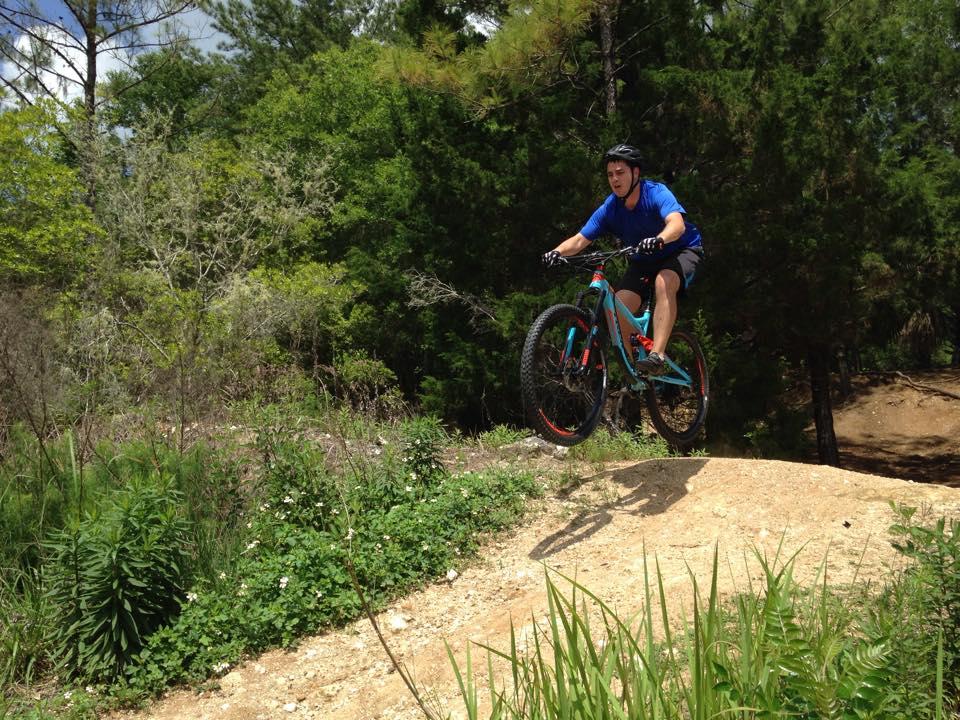 A person in a blue shirt and helmet is riding a mountain bike off a dirt ramp, airborne above a dirt trail surrounded by greenery and trees. Santos mountain bike trail.