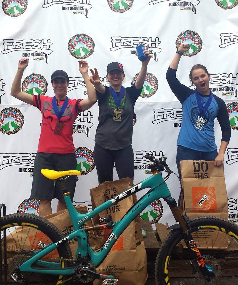 Three women stand proudly on a podium, each holding medals and celebrating their achievements. They are smiling and raising their arms in victory, with a teal mountain bike positioned in front of them. The backdrop features a branded banner for "FRESH Bike Service," and they are surrounded by brown paper bags with motivational phrases. Big Creek mountain bike trail.