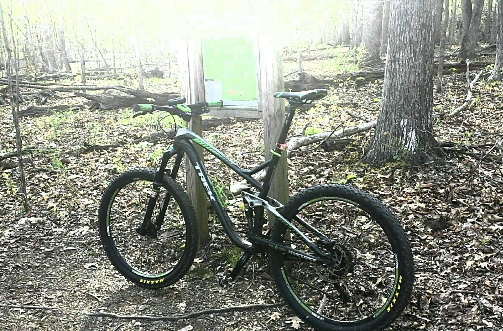 A mountain bike with a black and green design is parked near a trail sign in a wooded area. The ground is covered with fallen leaves, and trees surround the scene, indicating a natural environment. The sunlight creates a bright effect in the background. Schaeffer Farms mountain bike trail.