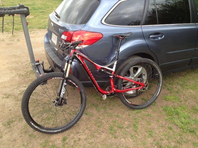 Specialized Stumpjumper FSR Comp 29er: A red mountain bike parked next to a blue SUV with a bike rack attached, set against a grassy background.
