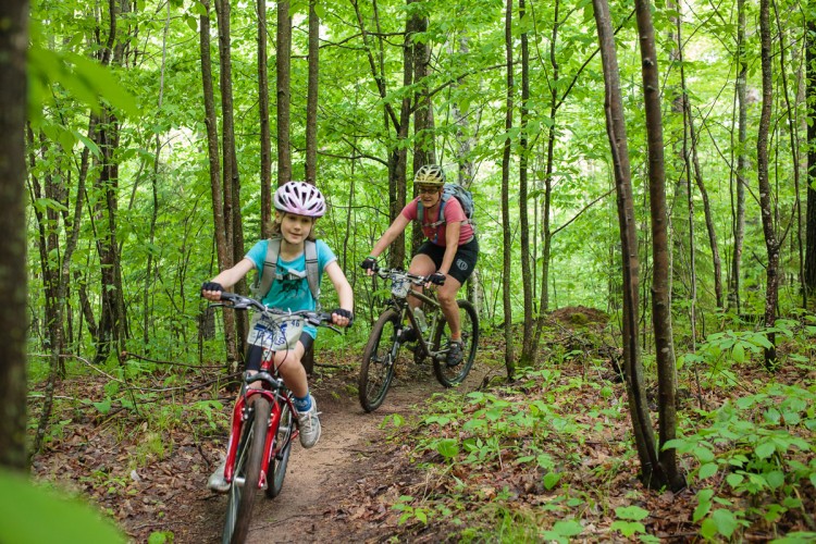 A child and an adult riding mountain bikes along a dirt trail in a lush green forest. The child, wearing a pink helmet and a blue shirt, is smiling as they pedal ahead. The adult follows closely behind, wearing a pink top and a helmet, both enjoying the tranquility of the natural surroundings with tall trees and greenery.