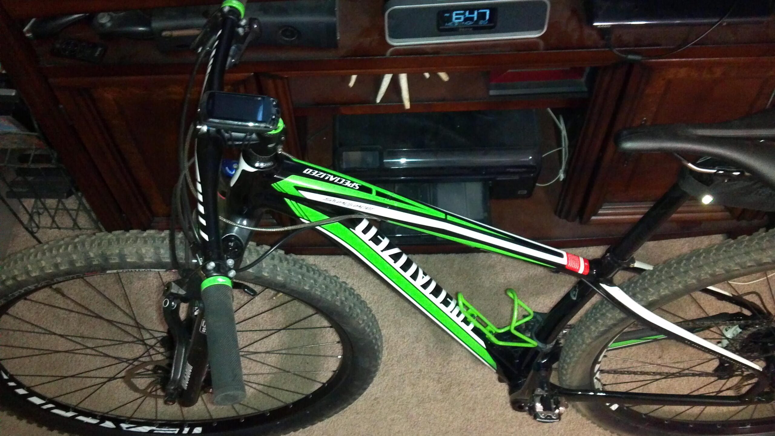 Specialized STUMPJUMPER COMP HT: A close-up view of a green and black mountain bike, featuring wide tires and a digital display mounted on the handlebars. The bike is positioned on a carpeted floor in a living room setting, with a wooden cabinet and electronic devices visible in the background.