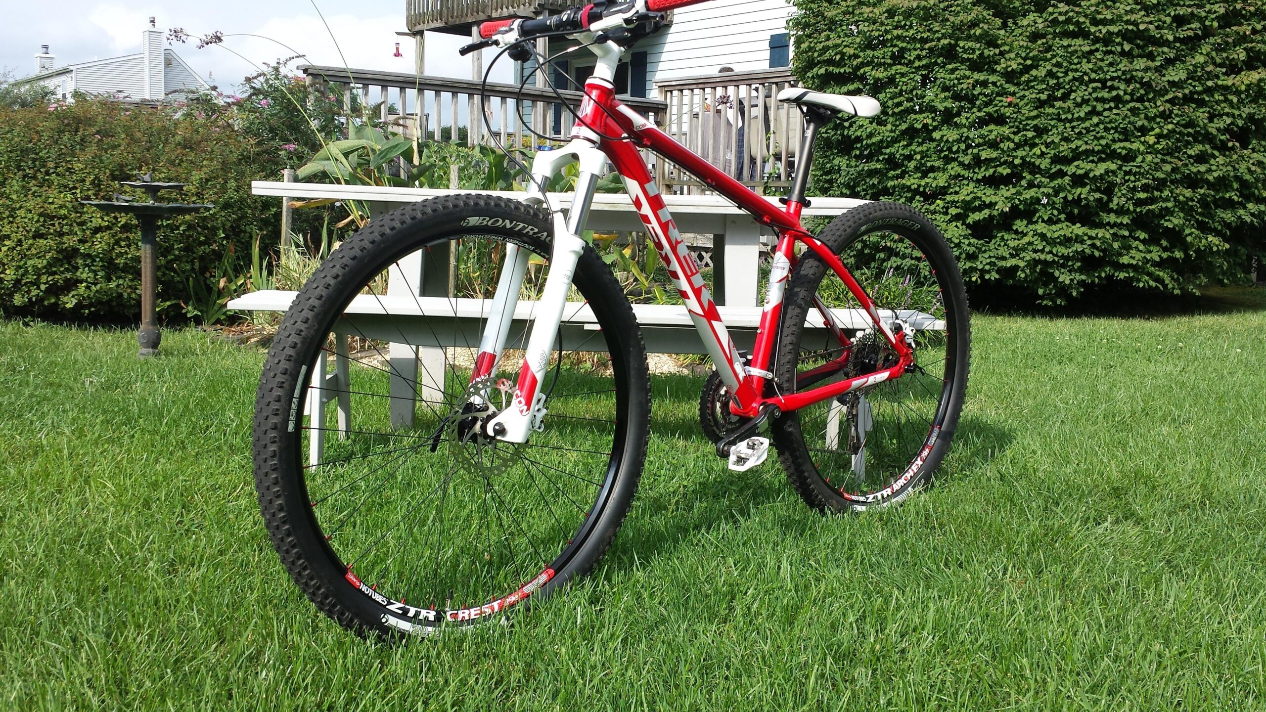 Trek Cobia: A red and white mountain bike is parked on a lush green lawn, with a wooden deck and shrubbery visible in the background. The bike features thick tires and a front suspension fork, showcasing its design for off-road riding.