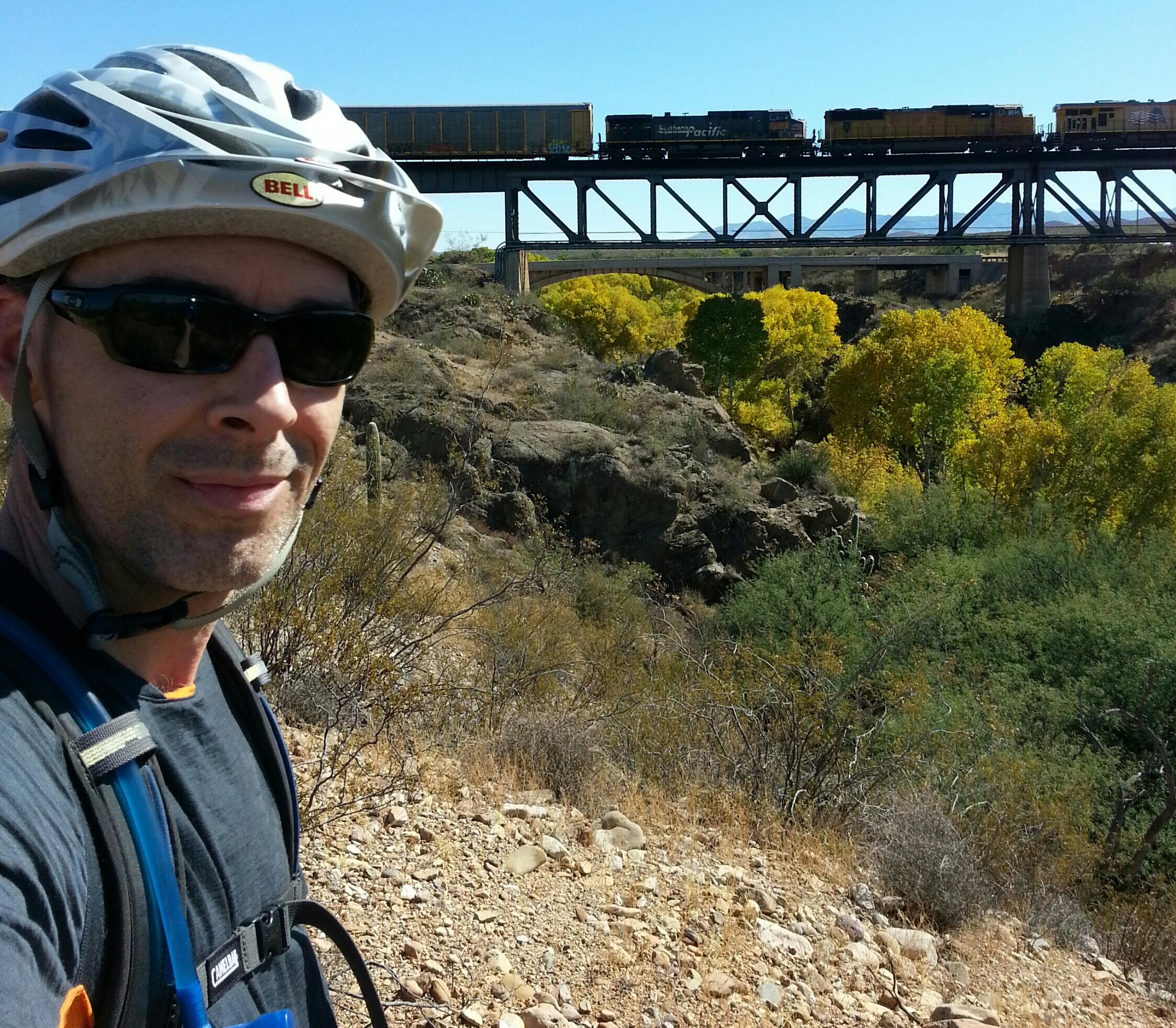 A cyclist wearing a white helmet and sunglasses stands in front of a rocky landscape with greenery and autumn-colored trees. In the background, a train crosses a bridge above, under a clear blue sky. Arizona Trail: Cienega Corridor mountain bike trail.