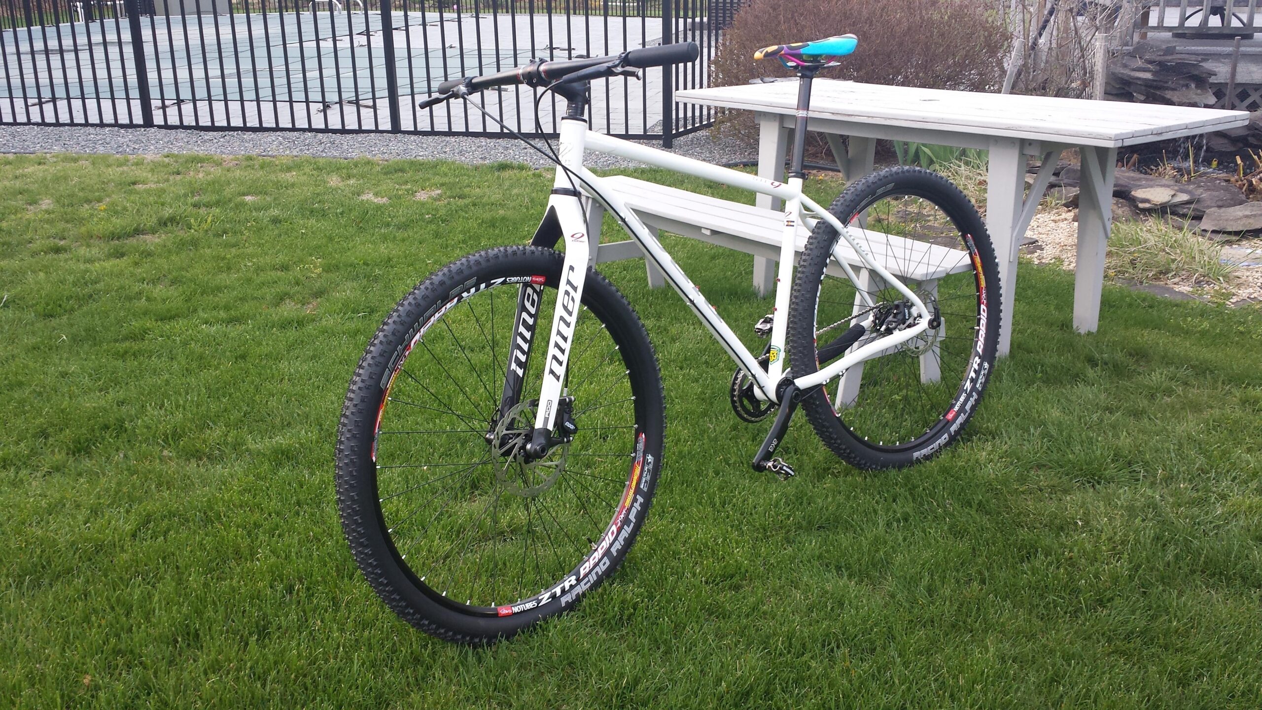 Niner S.I.R. 9: A white mountain bike with wider tires and a sleek frame, parked on green grass next to a wooden picnic table. In the background, there is a fenced area with a tennis court. The bike features branded rims and handlebars.