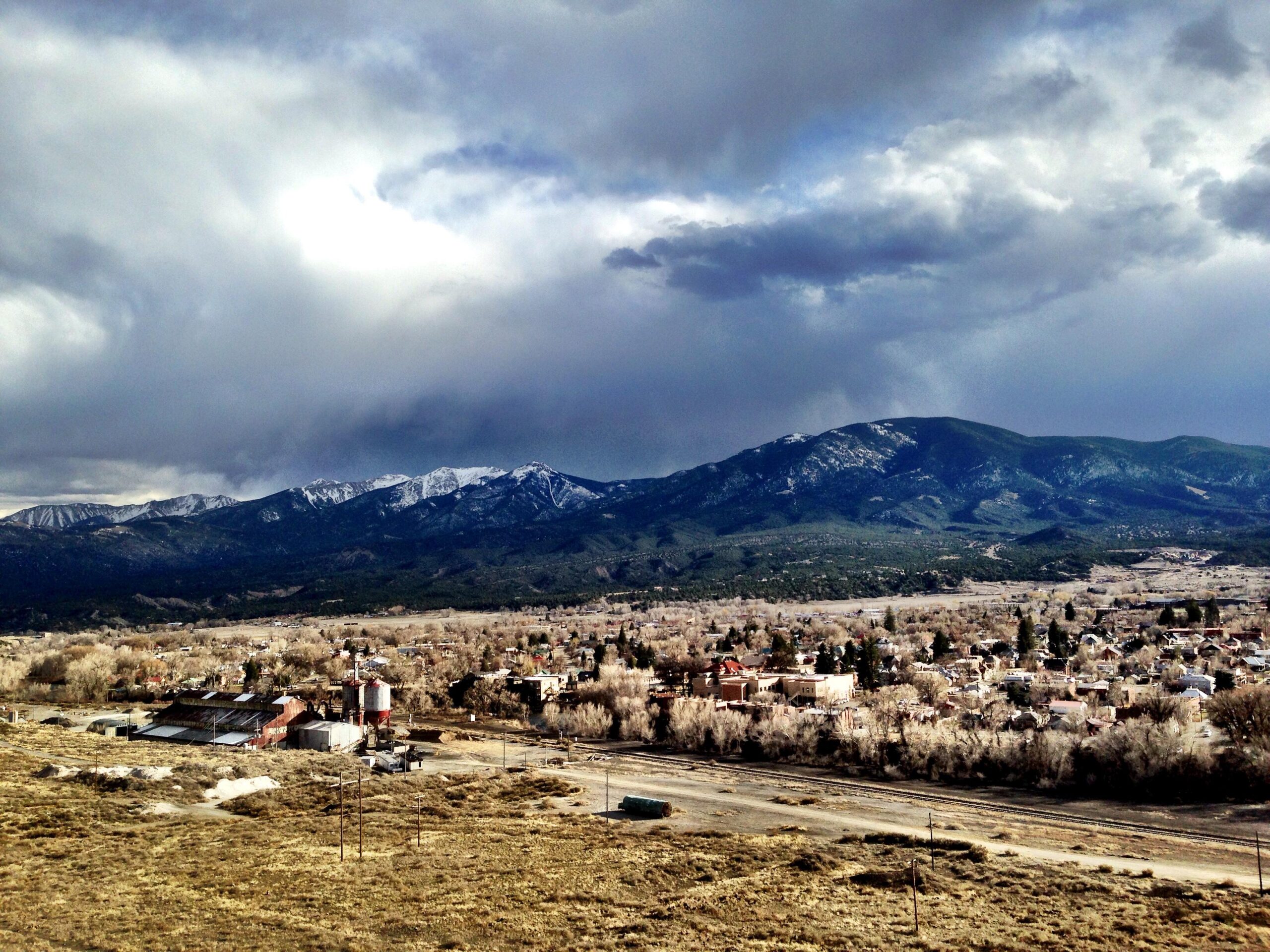 A panoramic view of a small town nestled in a valley, surrounded by mountains and trees. Snow-capped peaks rise in the background beneath a cloudy sky, while the foreground features a mix of residential buildings and industrial structures, with fields and a dirt road leading through the landscape. Arkansas Hills mountain bike trail.