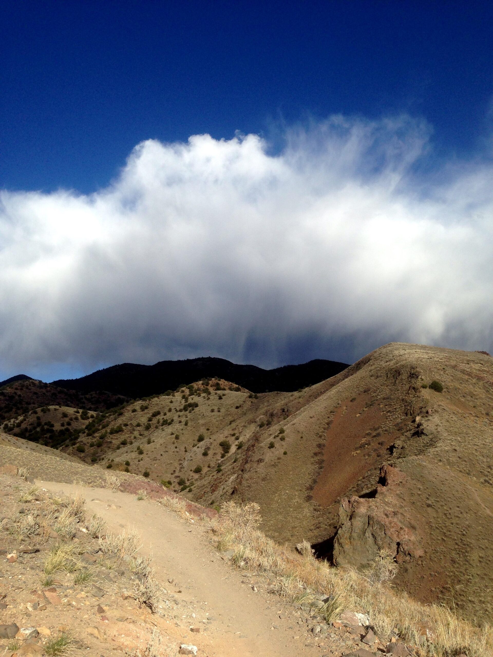 A winding dirt path leads through arid hills, with layered mountain ridges and patches of greenery in the foreground. Above, a dramatic sky features white clouds contrasted against a deep blue background. Arkansas Hills mountain bike trail.
