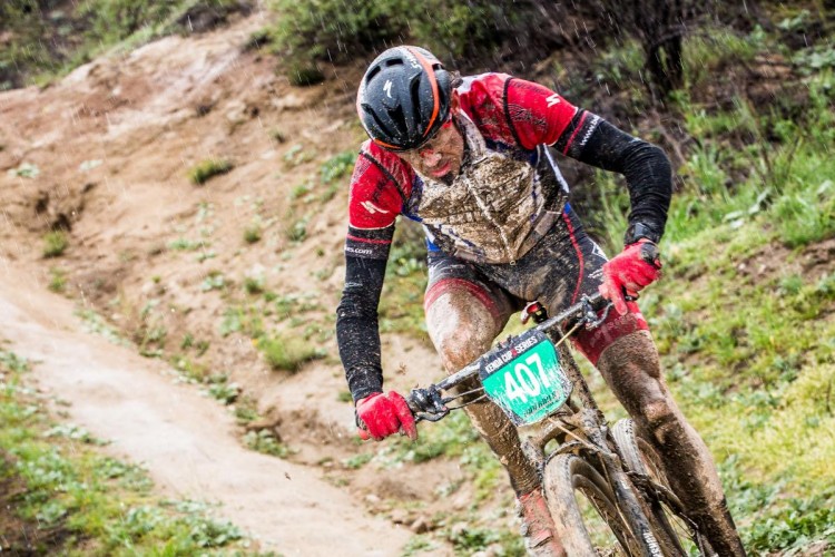 A muddy mountain biker competes in a rain-soaked race, intensely focused as he navigates a dirt pathway. His clothing is splattered with mud, and he wears a helmet and gloves, with a race number visible on his bike. The background features a blurred landscape of greenery and wet terrain.