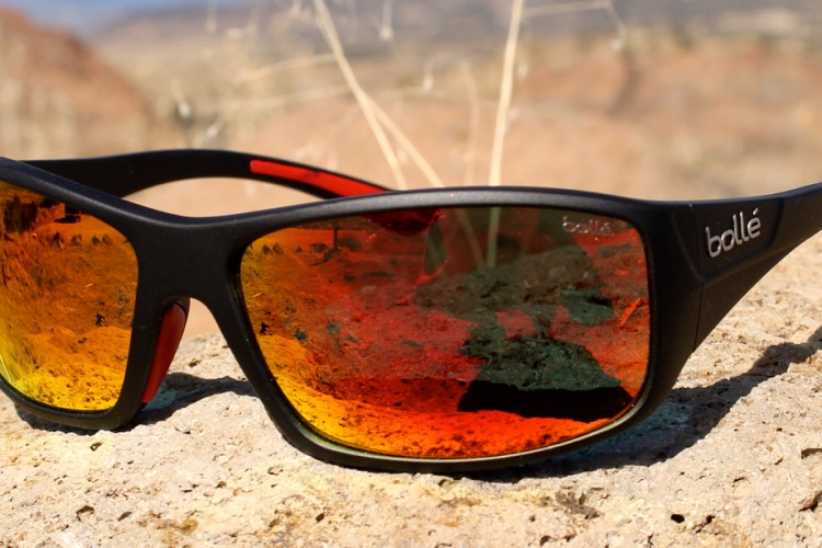 A pair of black sunglasses with reflective orange lenses resting on a rocky surface, set against a blurred outdoor background.