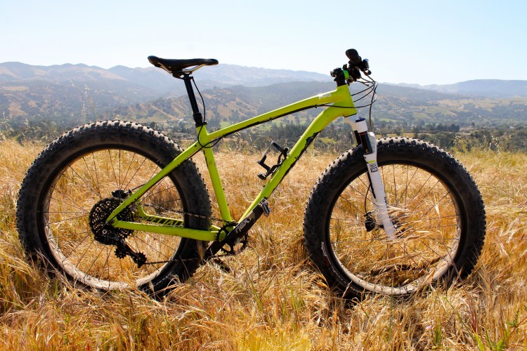 A bright green fat bike with oversized tires is positioned on a grassy hillside, showcasing a scenic view of rolling hills and mountains in the background under a clear blue sky.