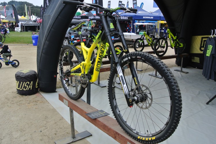 A brightly colored mountain bike displayed at a cycling event, featuring a yellow frame and thick tires. The bike is mounted on a wooden stand, with various bikes and vendor tents visible in the background. A child rides a small bike nearby, set on a sandy ground.
