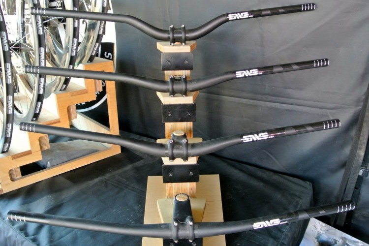 A display showcasing various black bicycle handlebars arranged on a wooden stand, with a blurred background of bicycle wheels and promotional materials. The handlebars have distinctive branding and design elements.